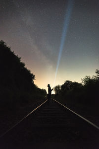 Silhouette person with illuminated flashlight standing on railroad track against sky at night