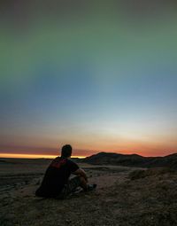 Rear view of man sitting on mountain by beach against cloudy sky during sunset