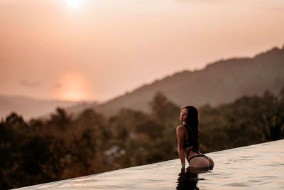 Rear view of woman sitting on at the edge of the pool against sky during sunset