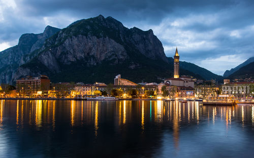 Illuminated buildings at waterfront against cloudy sky