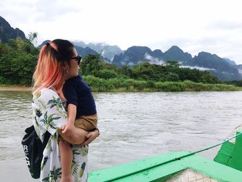 Mother carrying son while standing on riverbank against sky