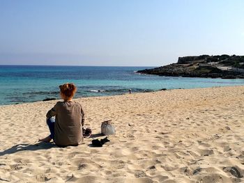 Rear view of man sitting on beach against clear sky