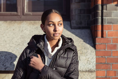 Thoughtful teenage girl wearing jacket looking away while sitting against wall