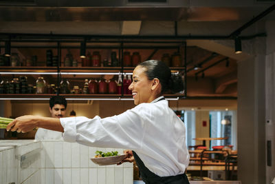 Happy female chef passing celery and holding salad bowl at commercial kitchen