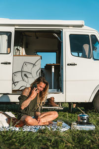 Full body of hipster female traveler having phone conversation while sitting on plaid with ukulele near camping van during road trip