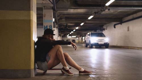 Side view portrait of woman sitting on skateboard in illuminated parking garage