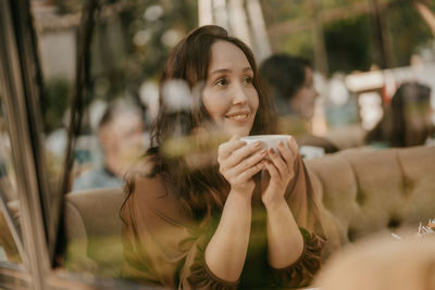 Smiling woman holding coffee at cafe seen through glass window