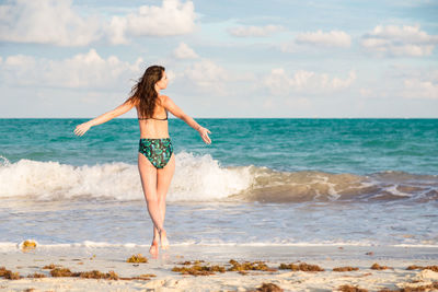 Rear view of woman wearing bikini while standing with arms outstretched at beach