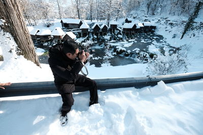 Man photographing while sitting on railing against river in forest during winter