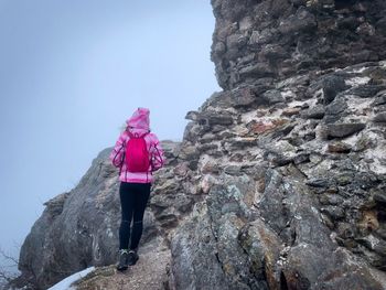 Woman with pink coat and backpack standing near tall cliffs surrounded by fog