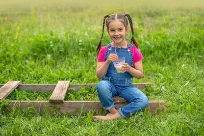 Funny girl in denim overalls and a pink t-shirt, holds a glass of milk on a green grass