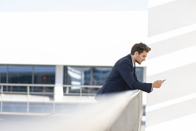 Businessman using smart phone while leaning on railing at office terrace