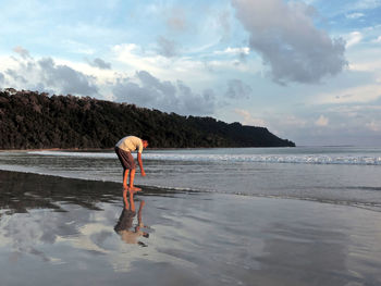 Man bending on shore by sea against cloudy sky