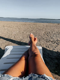 Low section of person relaxing on sea shore against sky