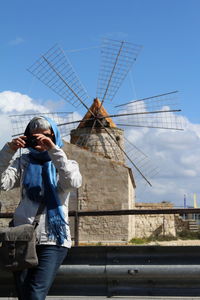 Woman in scarf photographing against traditional windmill against sky on sunny day