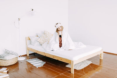 A young sick woman in pink pajamas and a sleeping mask on her head is sitting on the bed