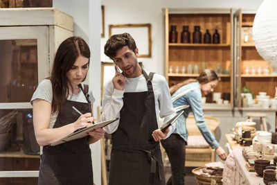 Male entrepreneur using phone while looking at female colleague writing on clipboard in boutique