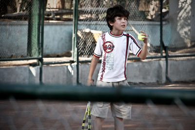 Portrait of  young boy standing against fence in the tennis playground 