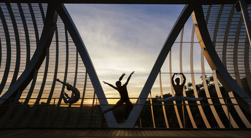 Woman with men dancing on footbridge against sky at sunset