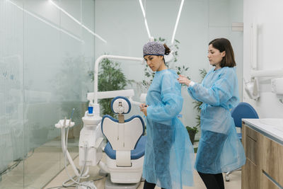 Female dentist helping assistant getting dressed in protective workwear at clinic