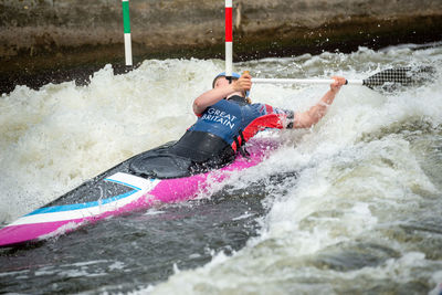 Gb canoe slalom athlete ducking under slalom pole while negotiating upstream gate in white water.