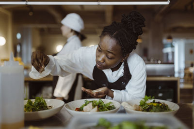 Focused young female chef sprinkling salt on salad while bending in commercial kitchen