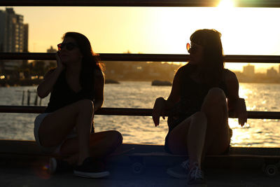 Friends sitting on skateboard against river during sunset