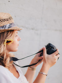 Side view of young woman wearing hat holding camera standing by wall outdoors