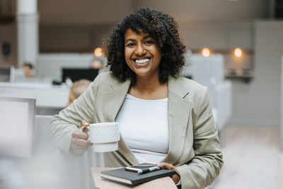 Happy female entrepreneur holding coffee cup and diary while standing in office