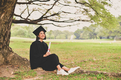 Smiling young woman in graduation gown holding certificate while sitting on grassy field at park