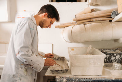 Side view of serious male artisan pouring liquid gypsum in wooden form for shaping material while creating artwork in workshop