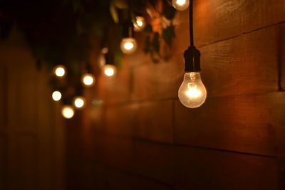 Close-up of illuminated light bulbs hanging from ceiling by wooden wall