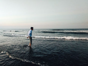 Side view of young man standing at beach against clear sky