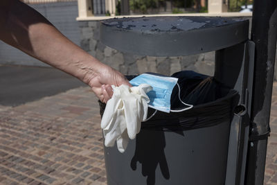 Cropped hand of person dumping glove and mask in garbage can