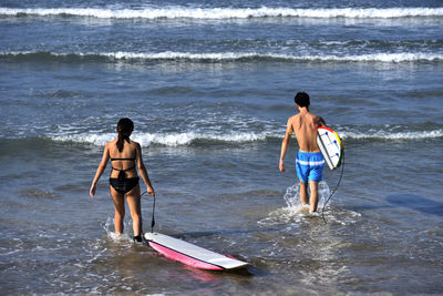 Rear view of friends holding surfboard while walking in sea