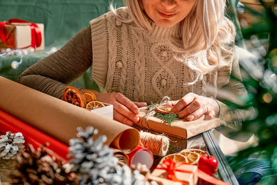 Blond woman wrapping presents in recycled card and decorated it with dried oranges and fir branches.