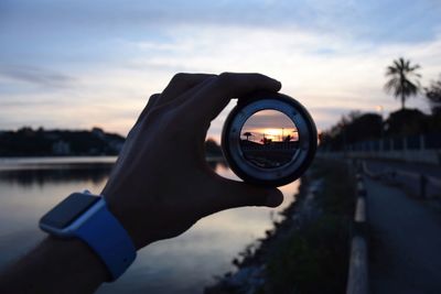 Close-up of hand holding magnifying glass against sea during sunset