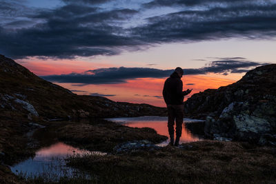 Man standing on rock by lake against sky during sunset