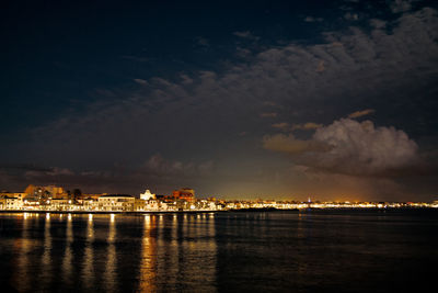 Illuminated buildings by sea against sky at night