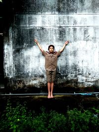 Full length of boy wearing police uniform while standing with arms raised against weathered wall