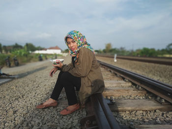 Full length side view portrait of woman sitting on railroad track against sky