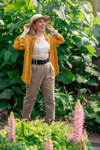 A girl in trousers and a shirt and a hat, stands in large green leaves, outside