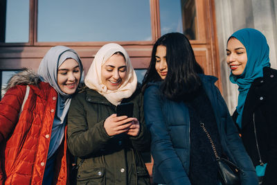 Happy female muslim friends sharing smart phone standing against entrance door in city