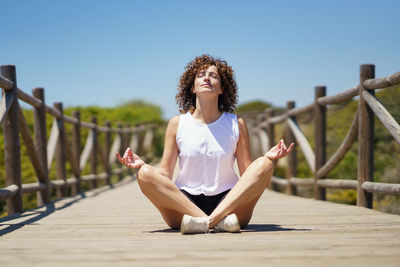 Portrait of young woman sitting on railing against clear sky