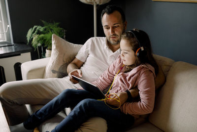 Father assisting daughter in using digital tablet on couch at home