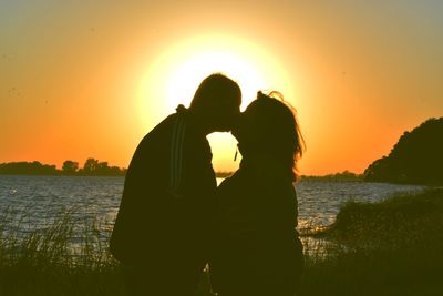 Silhouette couple kissing at lakeshore against clear orange sky