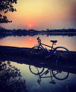 Bicycle parked by lake against sky during sunset