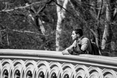 Thoughtful man looking away while standing by railing against bare trees