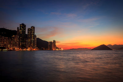 Sea by illuminated buildings against sky during sunset