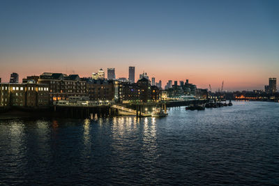 Illuminated buildings by river against sky at dusk